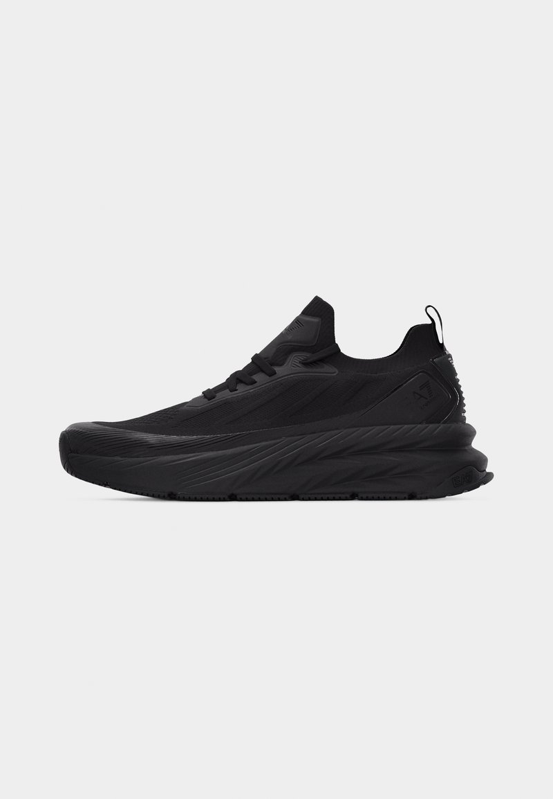 Baskets de sport noires avec semelle épaisse et texturée, design bas, lacets à l'avant et languette au talon, montrées de profil sur fond blanc.