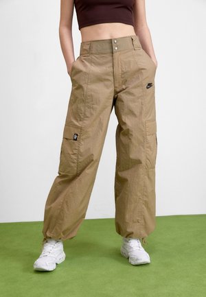 Osoba ubrana w beżowe spodnie typu cargo z logo Nike, krótki brązowy top, białe buty sportowe, stojąca na zielonej podłodze na tle białej ściany.