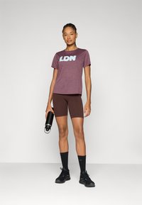 Camiseta marrom com gráfico "LDN", combinada com calções de bicicleta castanhos. Ténis e meias pretas, segurando uma garrafa de água preta. Fundo minimalista.