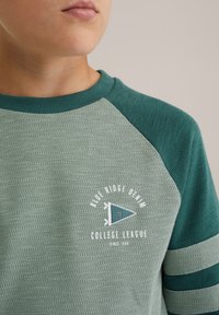 Közelkép egy gyermeki arcról, aki zöld, texturált pulóverben van, amelyen a "Blue Ridge Denim College League" logó és egy zászló látható a mellkasán.