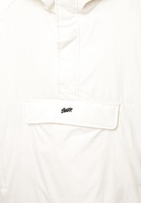 Weißer Blazer mit hohem Kragen, ausgestattet mit einer Fronttasche mit Klappe und einem gesteppten Design. Schwarzes Logo auf der Tasche; glatte Stofftextur.