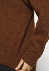 Main ajustant le poignet d’un pull brun pelucheux porté sur un pantalon foncé sur fond blanc.