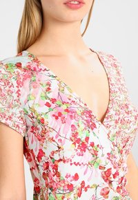 Kwiatowa sukienka w stylu wrap z lekkiego materiału charakteryzuje się głębokim dekoltem w kształcie V, krótkimi rękawami oraz kolorowym wzorem różowych i zielonych kwiatów.