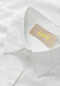 Hvid skjorte med krave, prydet med spredte sølvfarvede rhinsten. Etiket læser "JJXX" i gult på en beige baggrund. Tekstureret bomuldsstof.