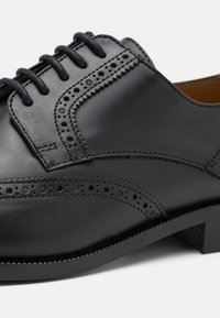 Une chaussure en cuir noir présente une finition lisse, des détails perforés décoratifs le long des bords et un embout rond avec un petit talon.
