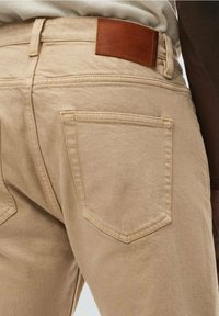 Pantalones de algodón beige con una textura suave, que presentan un parche de cuero marrón en la cinturilla y dos bolsillos traseros con bordes en ángulo.