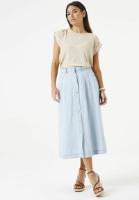 Hellblauer Jeansrock mit durchgängiger Knopfleiste, hoher Taille und fließender Silhouette. Kombiniert mit einem beigen Kurzarmoberteil und braunen Sandalen.