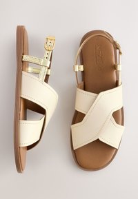 Sandales beiges avec un design à croisillons en matériau synthétique. Accents dorés sur les brides et la boucle. Semelle intérieure marron texturée. Semelle plate.