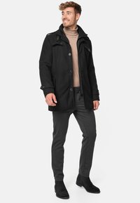 Musta villasekoitetakki, jossa on vetoketju- ja nappikiinnitys edessä, kaksi sivutaskua ja olkatoppaukset; yhdistetty harmaisiin housuihin ja mustiin saappaisiin.