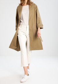 Beige, lehký trenčkot s širokými rukávy, délka ke kolenům a velkými bočními kapsami, kombinovaný s bílými zkrácenými kalhotami a teniskami.