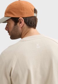 Sweat-shirt beige avec des manches raglan, texture douce et logo blanc à l'arrière ; casquette orange avec visière grise et sangle réglable.