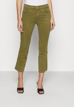 Pantalon olive fuselé et court avec ourlets effilochés et poches avant, porté avec des talons ouverts noirs et un haut blanc.