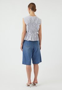 Haut léger à rayures avec une taille froncée associé à un short en jean bleu mi-cuisse. Le modèle porte des mules à talons blancs. Fond uni.