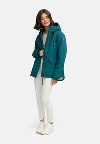Teal parka-jacka med huva, framfickor och trädetaljer. Buren över en grå topp, i kombination med vita jeans och sneakers.
