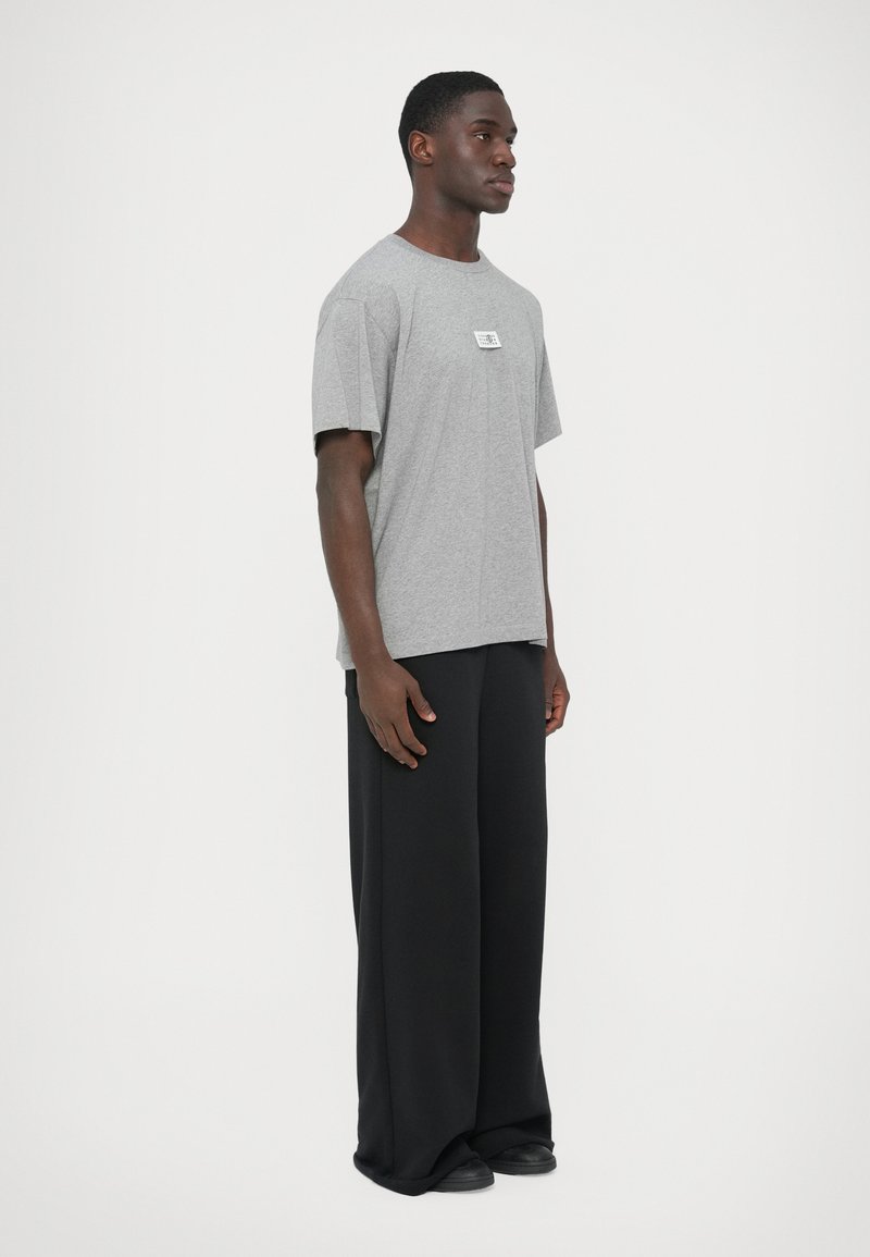 Gray cotton t-shirt with a crew neckline and a small logo patch, paired with loose-fitting black trousers, worn with black shoes.