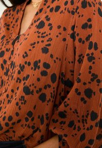 Brown blouse with a textured pattern of black spots, featuring a buttoned front and loose sleeves. The material appears lightweight and flowy.
