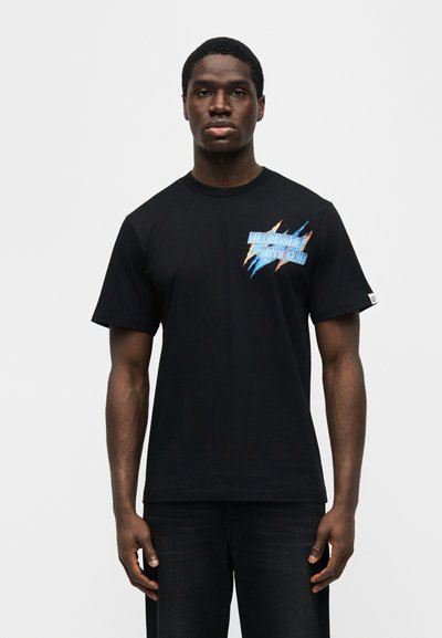 Νεαρός άνδρας που φοράει μαύρο T-shirt με λογότυπο "Billionaire Boys Club" σε μπλε και πορτοκαλί στον θώρακα, μπροστά σε απλό φόντο.