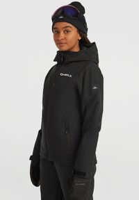 Zwarte waterdichte jas met capuchon, voorzien van een rits aan de voorkant, logo op de borst en elastische manchetten. Draagt een paar zwarte handschoenen en een beanie.