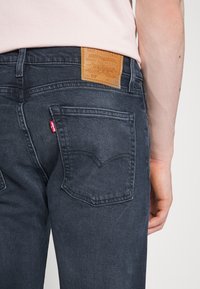 Vaqueros de mezclilla gris oscuro con un parche de cuero de la marca, diseño de cinco bolsillos y etiqueta roja contrastante en el bolsillo trasero, caracterizando una forma clásica.