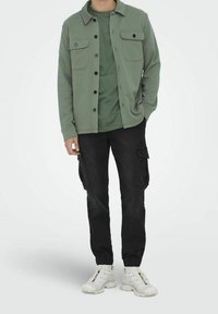 Chaqueta verde con botones y dos bolsillos en el pecho, llevada sobre una camiseta a juego, combinada con pantalones cargo negros y zapatillas blancas.