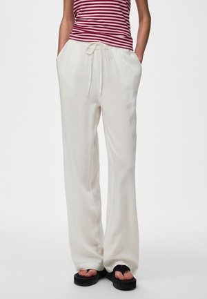 Kobieta ubrana w luźne białe spodnie ze sznurkiem, czerwono-biały pasiasty top bez rękawów oraz czarne klapki na platformie, ręce w kieszeniach.