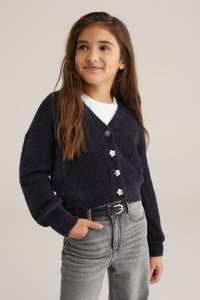 Mornarsko puhaste jopico z V-izrezom, petimi gumbi v obliki cvetov, oblečeno čez belo srajco, v kombinaciji s sivimi jeans hlačami.