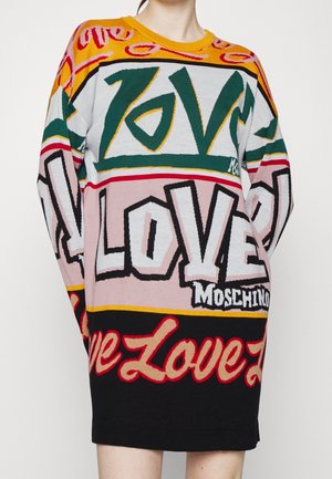 Osoba nosząca kolorową sukienkę sweterkową z długim rękawem z dużym tekstem "LOVE" i logo "Moschino" pogrubioną czcionką, mieszanymi wzorami i kolorami.