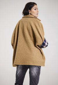Oversized tan fleece jacket with a textured finish and plaid cuffs. Pairing with dark denim jeans, showcasing a relaxed fit.