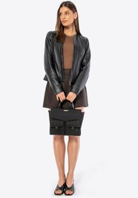 Black fabric handbag with two front pockets and gold hardware, held by a person wearing a black leather jacket and brown skirt.