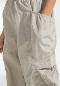 Letvægts, beige bukser med elastisk talje, sidelommer og mesh-detaljer på den nederste del, med en glat, krøllet tekstur.