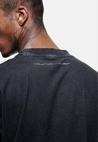 Zwarte katoenen T-shirt met een ronde hals en een subtiel handtekeningdetail op de achterkant. Gladde textuur met een relaxte pasvorm.