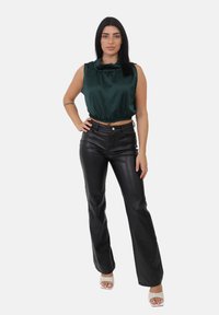 Kobieta z czarnymi włosami, nosząca ciemnozielony bezrękawnik satynowy, czarne skórzane spodnie i białe sandały na obcasie, stoi w pozie z jedną ręką na biodrze.