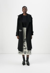 Black wool trench coat with wide lapels and a silver buckle detail, paired with a pleated skirt featuring a black floral print and heeled shoes.
