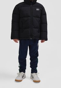 Personne portant une doudoune noire, un pantalon de survêtement bleu marine avec un logo, et des baskets blanches à semelles en gomme, debout devant un fond gris.