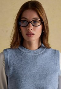 Noor, kellel on sirged kastanpruunid juuksed, kannab ümmargusi kilpkonnakujulisi prille ning helesinist ilma varrukateta kudumit, mille all on valge pikkade varrukatega särk.