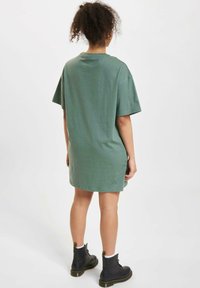 Przestronna zielona sukienka t-shirt z bawełny, z krótkimi rękawami, zaokrąglonym dolnym wykończeniem i bocznymi rozcięciami, zestawiona z czarnymi sznurowanymi botkami za kostkę.