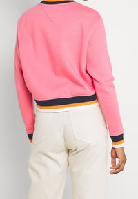 Růžová krátká mikina s námořnicky modrým a oranžovým žebrovaným lemem a manžetami, s klasickým kulatým výstřihem a hladkým, měkkým materiálem.