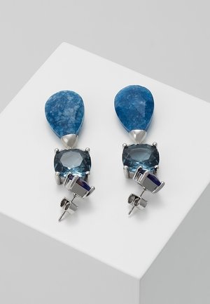 Silver earrings with a teardrop blue stone on top and a square blue gemstone below, resting on a white angled surface.