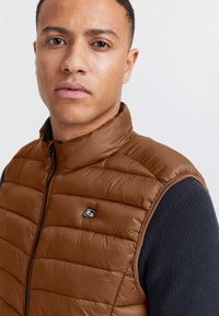 Braune Steppweste mit hohem Kragen, mit gesteppte Abschnitte und einem schwarzen Reißverschluss. Wird über einem dunkelblauen Pullover getragen. Logo auf der Brust.