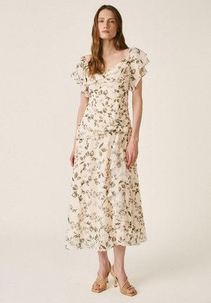 Mujer con vestido midi floral color crema con mangas con volantes y dobladillo asimétrico, combinando con sandalias beige de tacón, posando frente a un fondo liso.