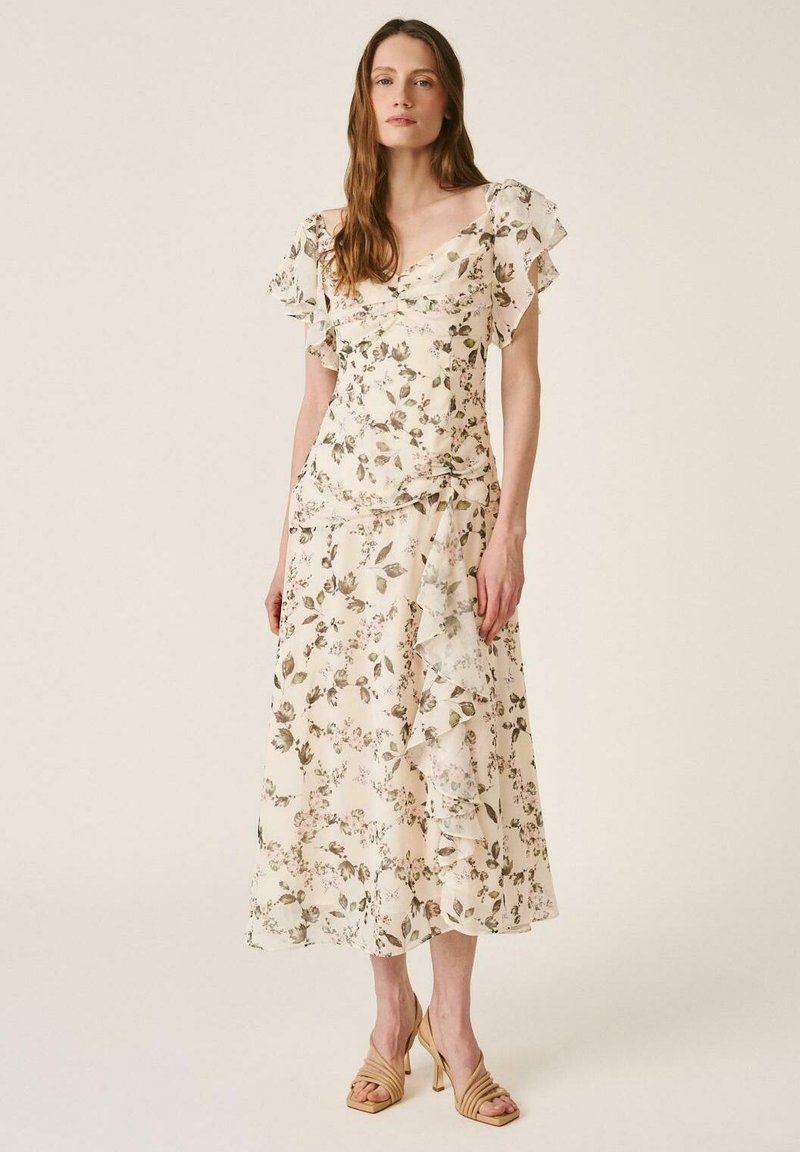 Mujer con vestido midi floral color crema con mangas con volantes y dobladillo asimétrico, combinando con sandalias beige de tacón, posando frente a un fondo liso.