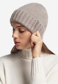 Jasnoszara dziergana czapka beanie z prążkowaną fakturą i zawiniętym brzegiem. Na boku znajduje się mała metka z nazwą marki.