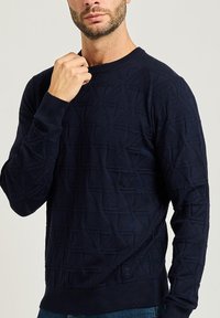 Maglione lavorato a maglia blu navy con un motivo geometrico testurizzato, collo a giro, e polsini a costine. Tessuto morbido con un design aderente.