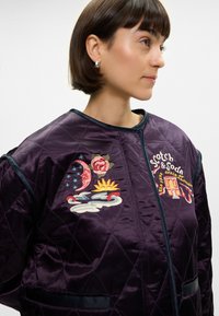 Κουκουλοφόρος μπομπέρ τζάκετ σε μοβ απόχρωση, με κεντημένα λουλουδάτα και ουράνια σχέδια, αντίθετο τιρκουάζ φινίρισμα και πλαϊνές τσέπες. Λείο σατέν υφασμά.