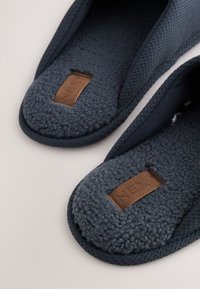 Sötétkék polár papucsok texturált szövet külsővel és barna, téglalap alakú márkajelzéssel, amely párnázott talpbetéten van varrva.