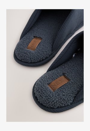 Dunkelblaue Fleece-Hausschuhe mit strukturiertem Stoff außen und braunen rechteckigen Markenschildern, die auf gepolsterten Einlegesohlen aufgenäht sind.