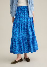 Gonna maxi con un design a strati, realizzata in tessuto blu con un motivo decorativo a spirale. Vita elasticizzata per il comfort, abbinata a scarpe basse.