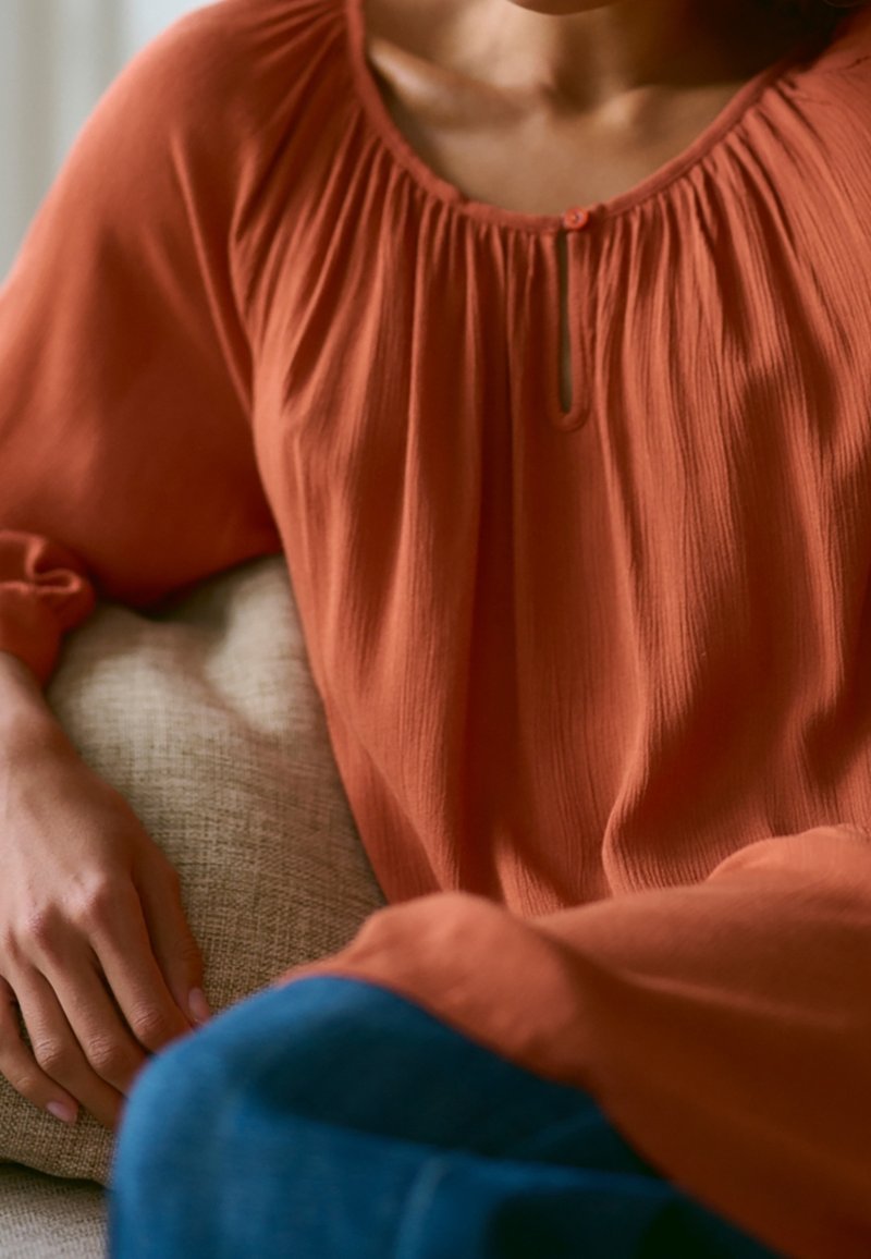 Personne portant une blouse orange ample avec un col froncé assise sur un canapé en tissu clair avec un pantalon bleu visible.