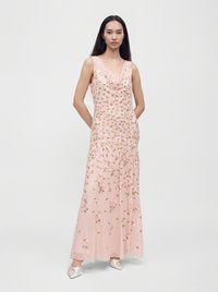 Femme aux longs cheveux foncés portant une robe maxi rose sans manches à fleurs et des chaussures blanches pointues, debout les mains derrière le dos.