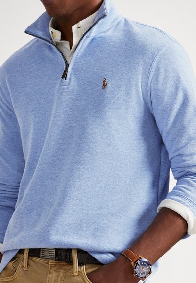 Vīrietis valkā gaiši zilu džemperi ar ceturtdaļas rāvējslēdzēju virs baltas krekliņa, brūnas krāsas bikses un rokas pulksteni ar brūnu ādas siksniņu un zilu ciparnīcu.